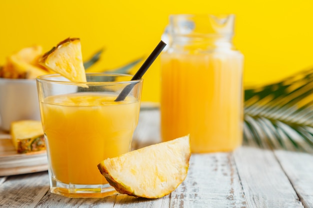 Jugo de piña sabroso en vaso con ingredientes. Cóctel de jugo de piña natural fresco y jugo de piña en jarra de vidrio sobre mesa de madera blanca con hojas de palma de fondo amarillo de verano.