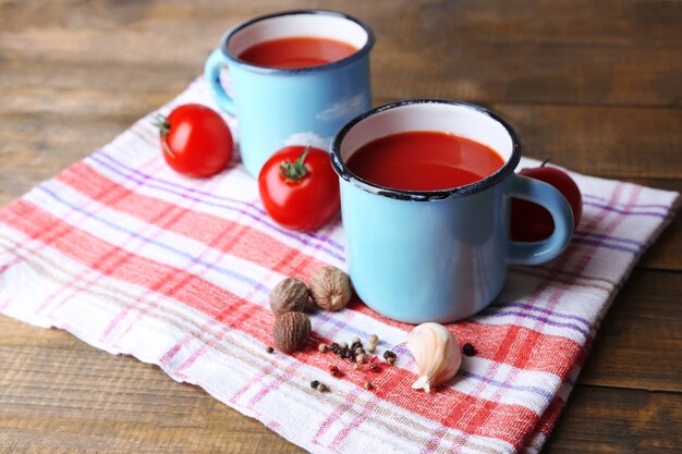 Jugo de tomate casero en tazas de colores, especias y tomates frescos en una servilleta, sobre fondo de madera