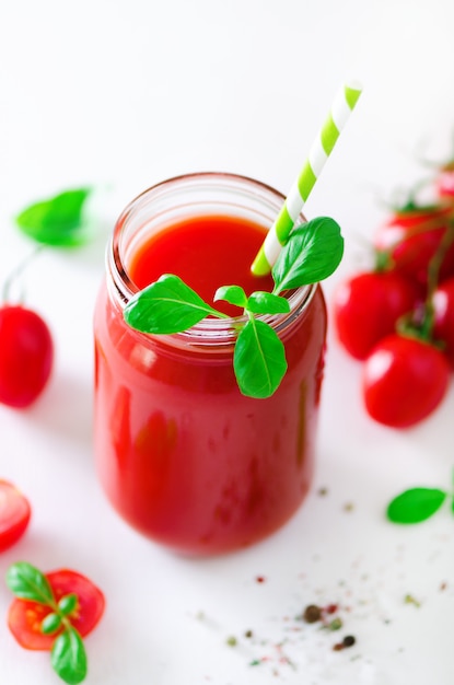 Jugo de tomate fresco orgánico en un frasco de vidrio, albahaca, cereza, sal, pimienta y paja a la luz. Concepto de alimentación y dieta limpia.