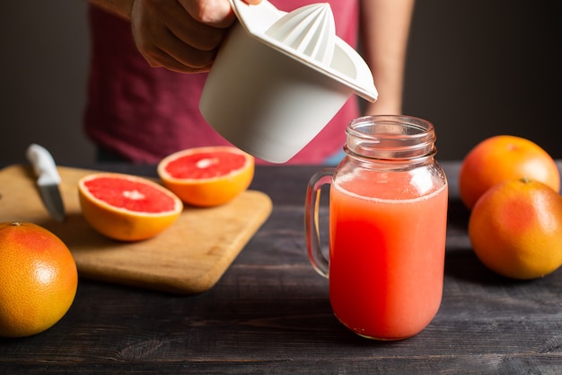 El jugo de toronja recién exprimido se vierte de un exprimidor manual en un frasco con asa. Exprimidor de plástico blanco en mano masculina. Sobre una mesa de madera negra hay toronjas enteras y en rodajas.