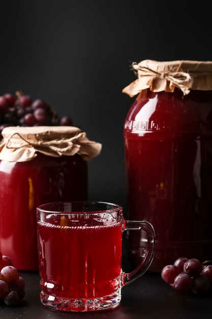 Jugo de uva en un vaso y latas, cocinado en un exprimidor, cosechando jugo de una cosecha de uva ubicada en una pared oscura, orientación vertical, primer plano