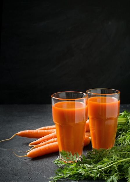 Jugo de zanahorias en vasos sobre fondo de piedra oscura con zanahorias frescas y crudas, copie el espacio, de cerca, vertical