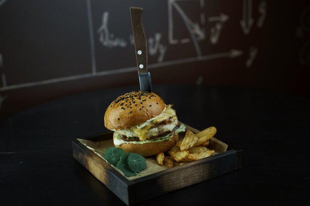 Jugosa hamburguesa con queso con cebolla morada con verduras con salsa César con pollo y rodajas de papas doradas al horno sobre una tabla de madera en un restaurante. Una ración de sabrosa hamburguesa