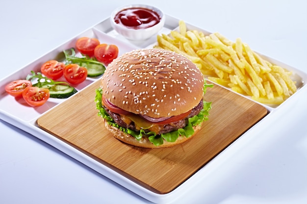 Jugosa sabrosa hamburguesa en una tabla de cortar de madera con papas fritas, verduras y salsa de tomate. Composición aislada sobre un fondo blanco y una bandeja de servir blanca.