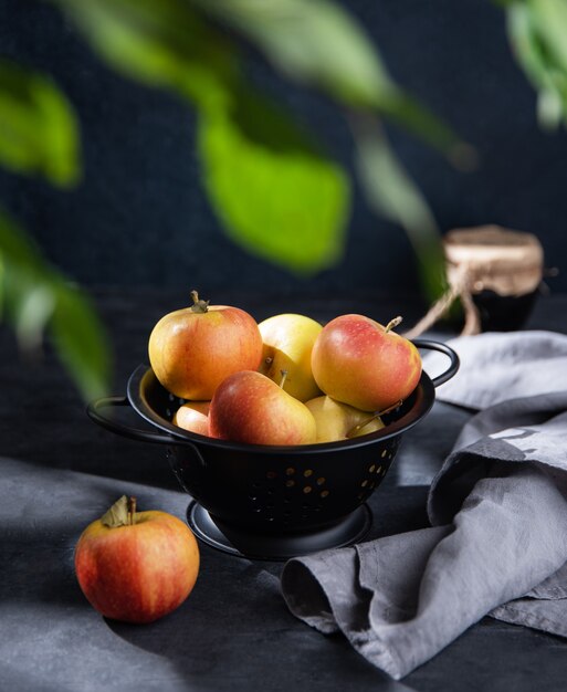 Jugosas manzanas en un recipiente negro con una toalla de lino y un tarro de mermelada sobre un fondo oscuro. Vista frontal e imagen oscura