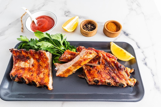 Jugosas rebanadas de costillas de cerdo a la parrilla en una bandeja oscura con verduras y salsa limonom y especias para mejorar el sabor de fondo blanco