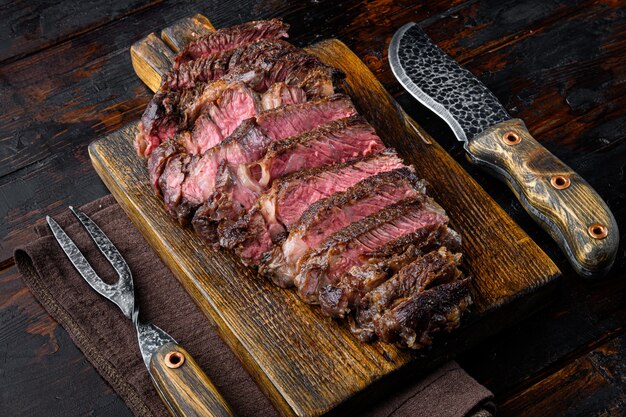 Jugosas rebanadas de filete Rib Eye de ternera mediana, sobre tabla de servir de madera, con cuchillo y tenedor de carne, sobre la vieja mesa de madera oscura