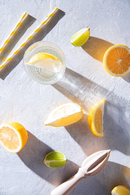 Jugosas rodajas de limón y lima y un vaso de agua mineral con limón sobre una mesa blanca con duras sombras matutinas. Vista superior