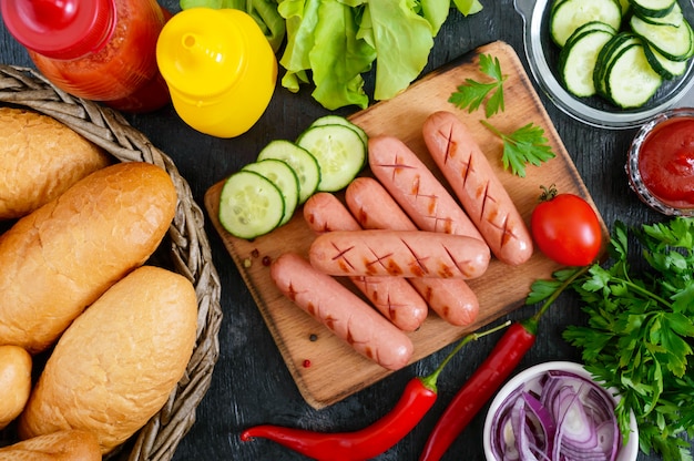 Jugosas salchichas a la parrilla, salsas, verduras frescas, bollos crujientes, sobre un fondo de madera. Vista superior. Flatlay Ingredientes para un hot dog. Comida de la calle.