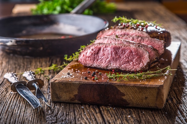 Jugoso medio filete Rib Eye de ternera en rodajas sobre placa de madera con tenedor y cuchillo hierbas especias y sal