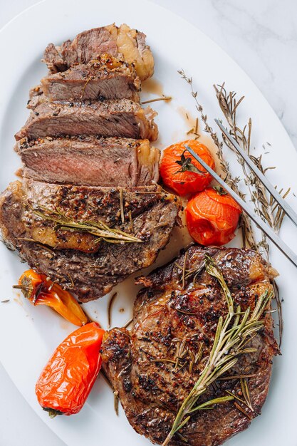 Jugosos filetes de chuletón bien fritos en un plato blanco sobre una superficie de mármol blanco con raspmarine y tomates cherry fritos y filetes de pimientos cortados en rodajas y listos para servir