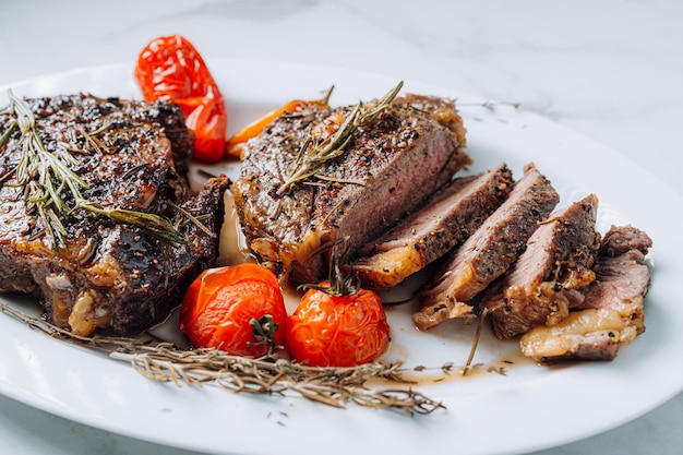 Jugosos filetes de chuletón bien fritos en un plato blanco sobre una superficie de mármol blanco con raspmarine y tomates cherry fritos y filetes de pimientos cortados en rodajas y listos para servir