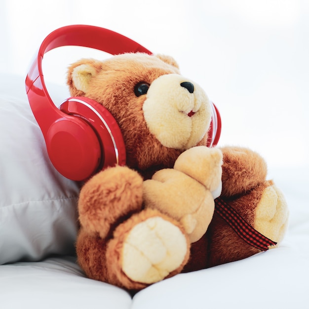 Juguete marrón encantador oso de peluche esponjoso que lleva creativamente auriculares rojos decorados en una cama romántica que se ve alegre con la música, causa deleite y relajación en unas felices vacaciones