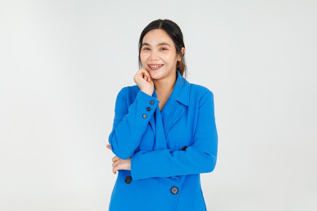 Juguetón y alegre niña asiática encantadora ríe y sonríe en el encantador traje de chaqueta azul, de pie con el brazo cruzado y la barbilla atrapada inocentemente. La hembra joven amistosa se ve agradable y adorable.