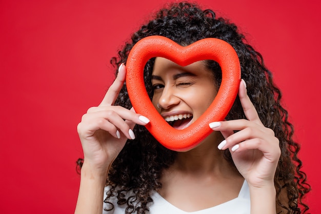 Juguetón guiño mujer negra mirando a través de la forma del corazón aislado en rojo