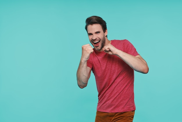 Juguetón joven sonriente en ropa casual gesticulando y sonriendo mientras está de pie contra la pared azul