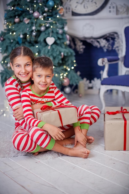 Juguetón sonriente feliz lindo niño y niña vestidos con pijama de rayas con regalo sentado en la habitación decorada de año nuevo en casa. Buen humor navideño. Concepto de estilo de vida, familia y vacaciones 2018