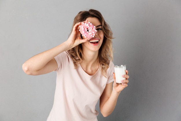 Juguetona mujer en camiseta divirtiéndose mientras sostiene donut y leche sobre gris