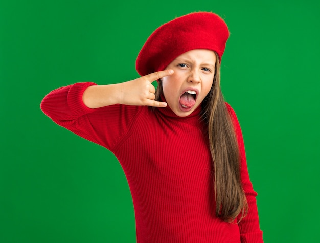 Juguetona niña rubia vistiendo boina roja haciendo signo de rock mostrando la lengua mirando al frente aislado en la pared verde