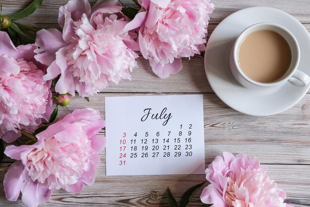 Juli 2022 Kalenderpfingstrosen und eine Tasse Kaffee auf dem Tisch Frauen-Desktop