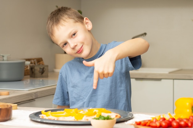 Junge 7-10 im T-Shirt, das Pizza in der Küche kocht, Kind lächelt und zeigt Finger auf Pizza