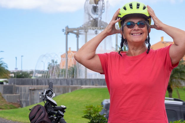 Junge ältere Frau in Aktivität mit Fahrrad im städtischen öffentlichen Park. Sie trägt einen gelben Schutzhelm. Brunnen im Hintergrund