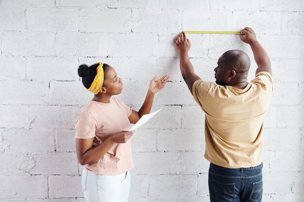 Junge Afrikanerin mit Papier, die neben ihrem Mann steht, die Länge der Mauerziegel misst und sich bei der Hausrenovierung mit ihm berät