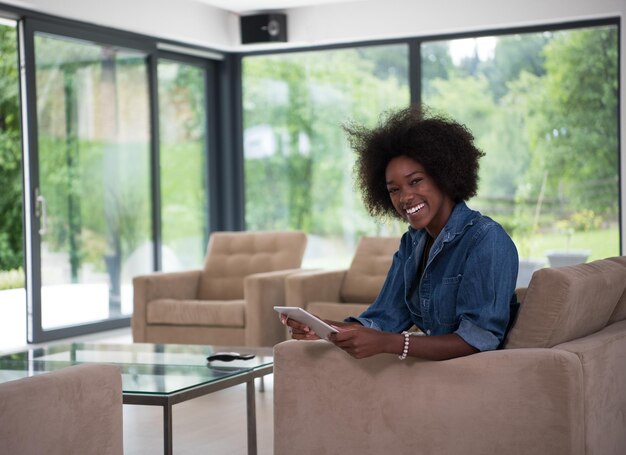 Junge afrikanisch-amerikanische Frau entspannt sich zu Hause in ihrem luxuriösen Wohnzimmer und liest einen digitalen Tablet-PC, surft im Internet und arbeitet