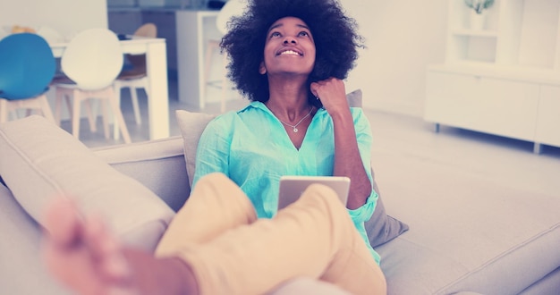 Junge afrikanisch-amerikanische Frau entspannt sich zu Hause in ihrem luxuriösen Wohnzimmer und liest einen digitalen Tablet-PC, surft im Internet und arbeitet