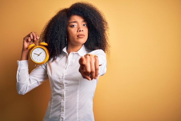 Junge afrikanisch-amerikanische Frau mit Afro-Haaren, die einen klassischen Wecker über gelbem Hintergrund hält und mit dem Finger auf die Kamera zeigt und auf Ihre Handzeichen positive und selbstbewusste Geste von vorne