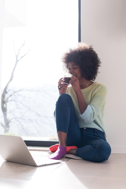 Junge afrikanisch-amerikanische Frau sitzt lächelnd auf dem Boden in der Nähe eines hellen Fensters, während sie auf einen offenen Laptop-Computer schaut und zu Hause eine Tasse hält
