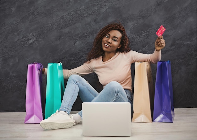 Junge afrikanische Frau, die zu Hause online einkaufen. Mädchen sitzt zwischen bunten Einkaufstüten