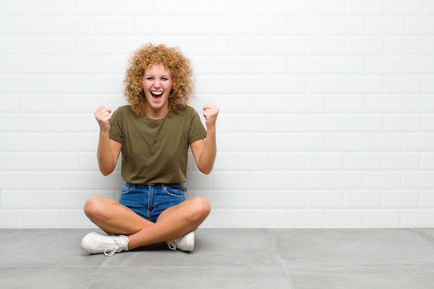 Junge Afro-Frau, die glücklich, überrascht und stolz fühlt, schreit und Erfolg mit einem großen Lächeln feiert, das auf einem Boden sitzt