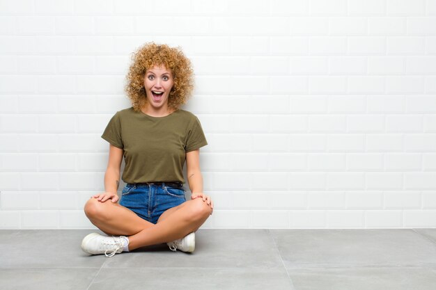 Junge Afro-Frau, die glücklich und angenehm überrascht aussieht, aufgeregt mit einem faszinierten und schockierten Ausdruck, der auf einem Boden sitzt