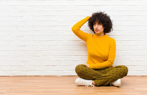 Junge Afro-Frau, die sich verwirrt und verwirrt fühlt, sich am Kopf kratzt und zur Seite schaut, die auf dem Boden sitzt