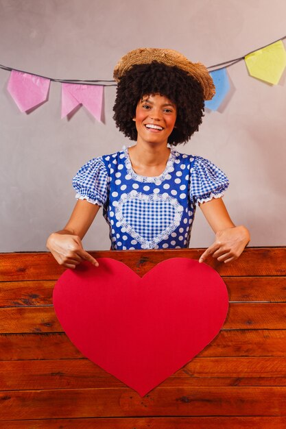 Junge Afro-Frau, gekleidet für Junina-Party hinter einem Holzbrett mit Herz