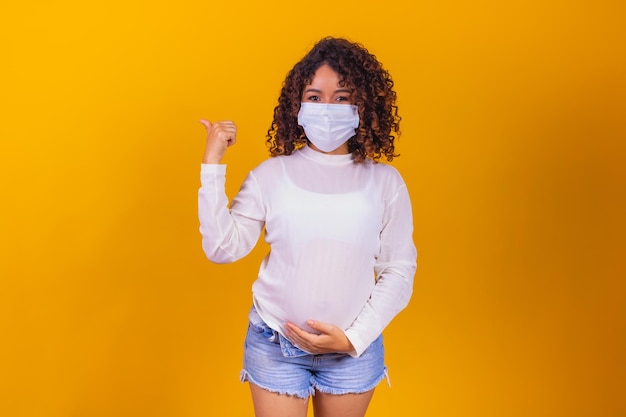 Junge afro-schwangere Frau mit chirurgischer Maske gegen Kovid, die auf freiem Platz für Text zur Seite zeigt. Schwangerschafts- und Krankheitspflegekonzept
