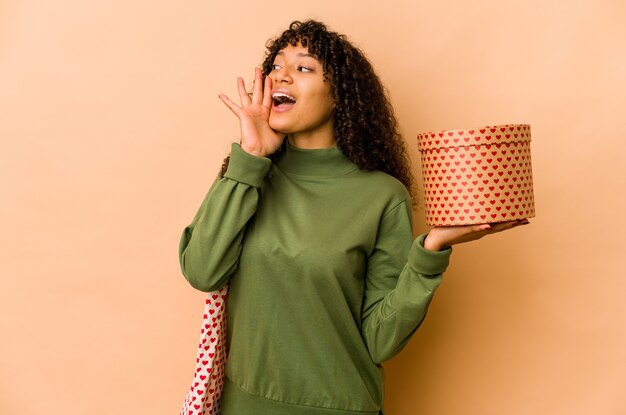 Junge Afroamerikaner-Afro-Frau, die ein Valentinstaggeschenk schreit und Handfläche nahe geöffnetem Mund hält.