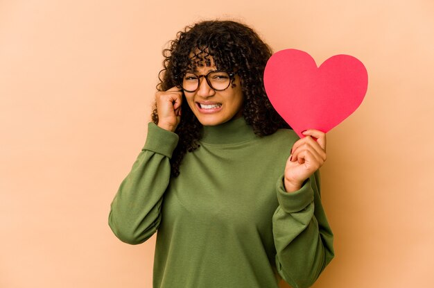 Junge Afroamerikaner-Afro-Frau, die ein Valentinstagherz hält, das Ohren mit Händen bedeckt.