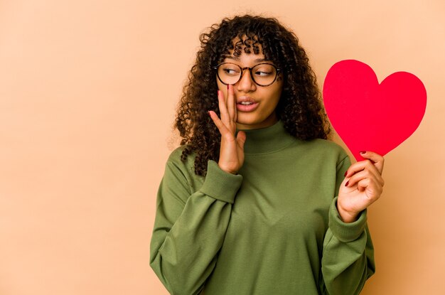 Junge Afroamerikaner-Afro-Frau, die ein Valentinstagherz hält, sagt eine geheime heiße Bremsnachricht und schaut zur Seite