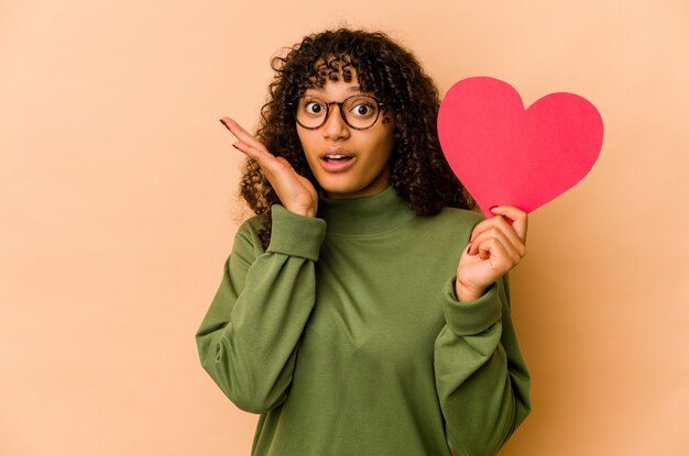 Junge Afroamerikaner-Afro-Frau, die ein Valentinstagherz überrascht und schockiert hält.