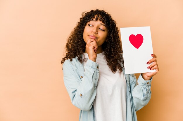 Junge Afroamerikaner-Afro-Frau, die eine Valentinstagskarte hält, die seitwärts mit zweifelhaftem und skeptischem Ausdruck schaut.