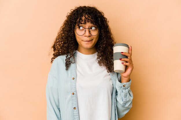 Junge Afroamerikaner-Afro-Frau, die einen Kaffee zum Mitnehmen verwirrt hält, fühlt sich zweifelhaft und unsicher.