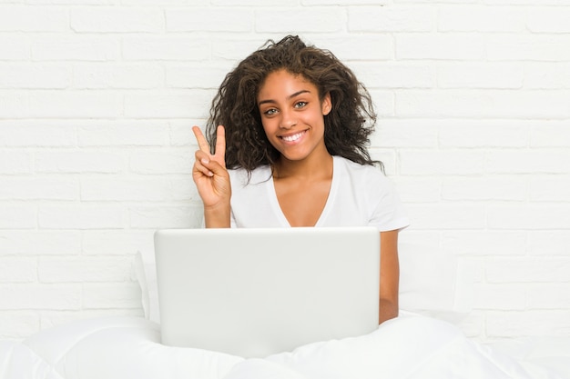 Junge Afroamerikanerfrau, die auf dem Bett mit Laptop sitzt und Nummer zwei mit den Fingern zeigt.