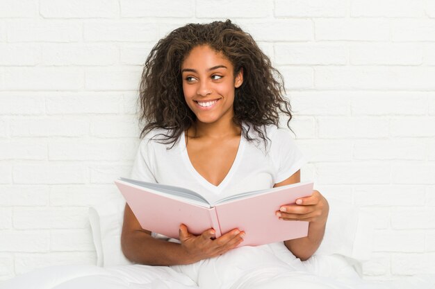 Junge Afroamerikanerfrau, die auf dem Bett studiert das Lächeln überzeugt mit den gekreuzten Armen sitzt.