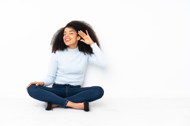 Junge Afroamerikanerfrau, die auf dem Boden sitzt und etwas hört, indem man Hand auf das Ohr legt