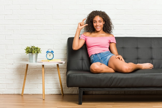 Junge Afroamerikanerfrau, die auf dem Sofa hält etwas wenig mit den Zeigefingern, lächelt und überzeugt sitzt.