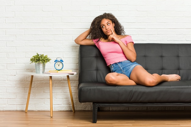 Junge Afroamerikanerfrau, die auf dem Sofa seitlich schaut mit zweifelhaftem und skeptischem Ausdruck sitzt.