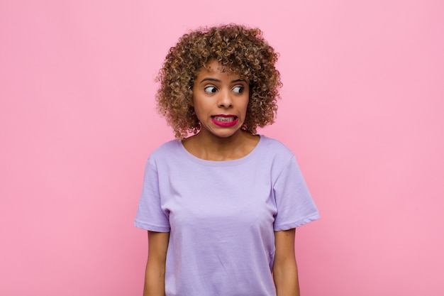 Junge Afroamerikanerfrau, die besorgt, gestresst, ängstlich und ängstlich schaut, in Panik gerät und Zähne gegen rosa Wand zusammenbeißt