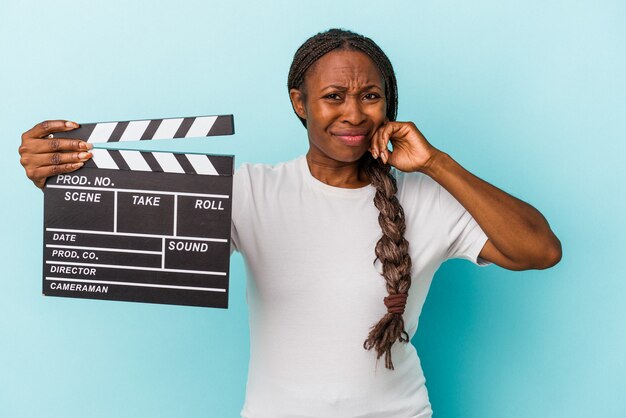 Junge Afroamerikanerfrau, die clapperboard lokalisiert auf blauem Hintergrund hält, der Ohren mit den Händen bedeckt.